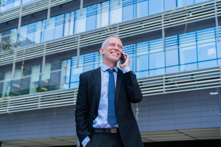 Confident businessman engaging in a phone conversation while smiling and dressed in a professional suit with a stylish tie in a modern corporate environment with a bright demeanorの写真素材