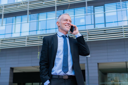 Confident businessman in a stylish suit engaging in a phone conversation while smiling and standing outdoors in a modern urban setting with a professional demeanor and polished appearanceの写真素材