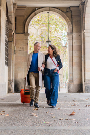 Happy couple enjoying a leisurely stroll together while holding hands and pulling a suitcase in a picturesque urban setting filled with beautiful architecture and vibrant colorsの写真素材