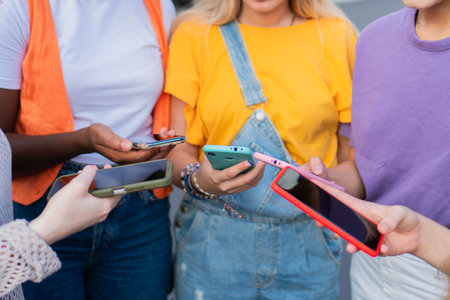 Group of diverse friends joyfully sharing their smartphones while engaging in lively conversation and exploring social media together in a vibrant, colorful setting filled with excitementの写真素材