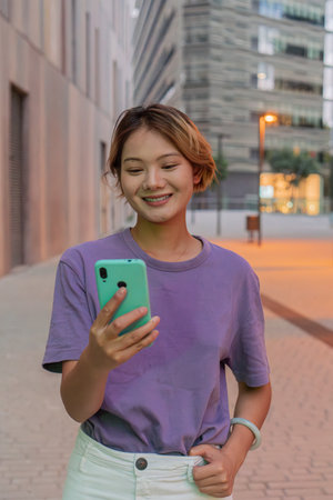 Smiling young woman wearing a purple shirt joyfully interacting with her smartphone while enjoying leisure time outdoors during a pleasant evening in an urban environmentの写真素材
