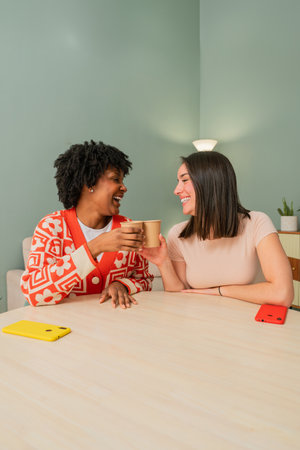 Two joyful women sharing a delightful moment while toasting with warm drinks and enjoying each others company during a friendly conversation at home in a cheerful atmosphere of camaraderieの写真素材