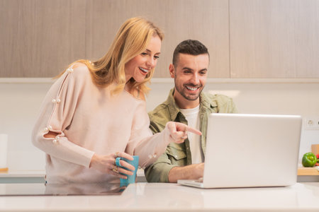 Joyful couple sharing exciting moments while engaging with a laptop in their cozy kitchen, filled with laughter and smiles during a delightful afternoon together, enjoying the time spent onlineの写真素材