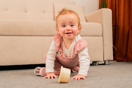 Happy baby crawling on soft carpet in cozy living room, playing with a toy while wearing adorable pink overalls and a white long-sleeve shirt, showcasing a joyful moment of childhood explorationの写真素材
