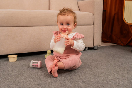 Cute and cheerful baby girl sitting on a soft gray carpet, happily enjoying a bottle of milk while surrounded by colorful toys in a cozy and modern living room settingの写真素材