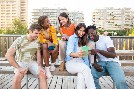 Group of young people standing together while sitting in wooden bench in the city - Funny multiethnic students watching their smartphones in the university campusの写真素材