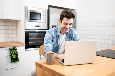 Handsome young man working with a laptop and smiling at the kitchen with a cup of tea - Cheerful, success and business conceptの写真素材