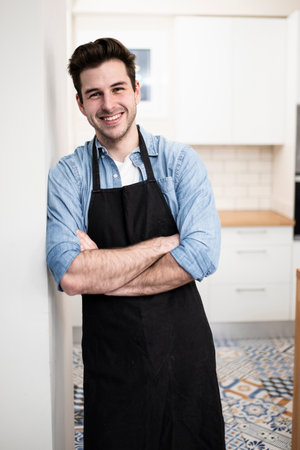 handsome young man smiling in the kitchen wearing an apron - Portrait of a cheerful confident caucasian young man looking at camera with arms crossedの写真素材