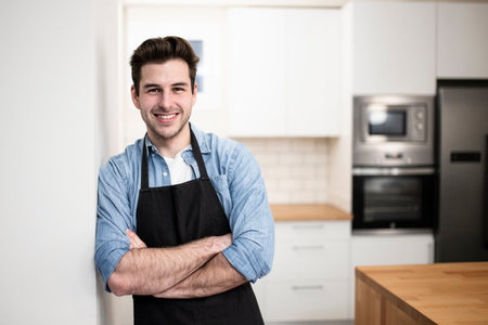 handsome young man smiling in the kitchen wearing an apron - Portrait of a cheerful confident caucasian young man looking at camera with arms crossedの写真素材