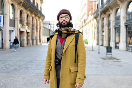 Young tourist with beard standing at the city looking at camera. Front view of a hipster male in...