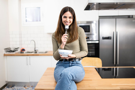 Portrait of beautiful young woman having breakfast in the kitchen. smiling female with a bowl ans spoon looking at camera. Lifestyle concept.の写真素材