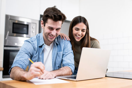 young caucasian couple planning budget together at home writting notes to paper and looking a laptop. two cheerful young adults sitting on the kitchen. finance conceptの写真素材