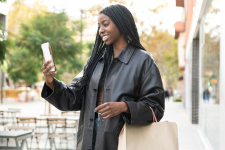 Happy young woman taking a videocall on phone in the street holding a shopping bag. Happy female using a smartphone in the streetの写真素材