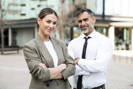 Confident and smiling business people looking at camera in the street with their arms crossed. Young businesswoman with a senior businessman behind looking satisfied.の写真素材
