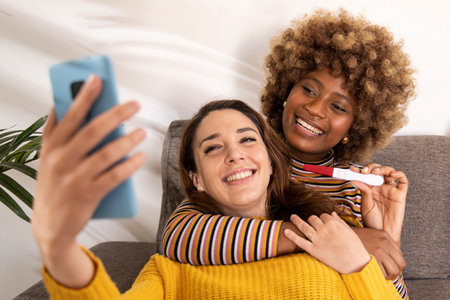 Diverse female couple taking a selfie with their positive pregnancy test in a sofa. Happy lesbian women announcing a pregnancy on their social media in a living room.の写真素材