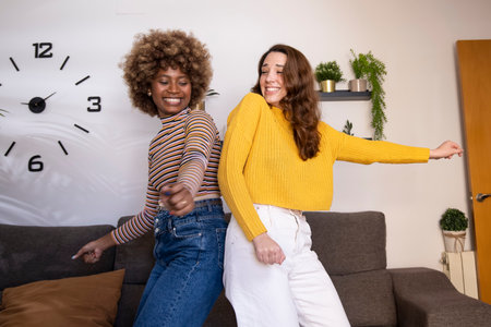 Multiracial young funny women dancing and laughing back to back in their apartment. Side view of happy and excited couple of girls celebrating in their living room.の写真素材