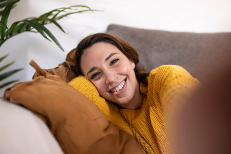 Close-up young cheerful woman looking at camera taking a selfie. Beautiful smiling female taking a selfie with smartphone while relaxing at home.の写真素材