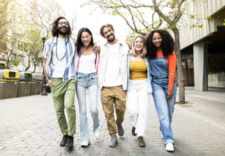 Group of cheerful millennial friends walking together hugging each other in city street. Happy...