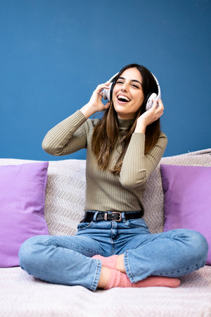 Cheerful young woman listening to music and smiling sitting on couch in livinig room. Happy young girl having fun at home.の写真素材
