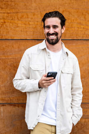 Confident handsome young guy texting with his phone and looking at camera. Happy and relaxed man using his smartphone standing on a brown wall outside staring at camera.の写真素材