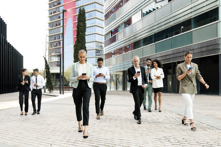 Group of confident business people walking the street using their phones. Diverse team of focused businesswoman and businessman walking outside while texting on their smartphones.の写真素材