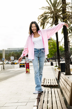 Young smiling woman balancing on bench in the street. Carefree and relaxed lady walking on bench park with her arms outstretched in a city.の写真素材