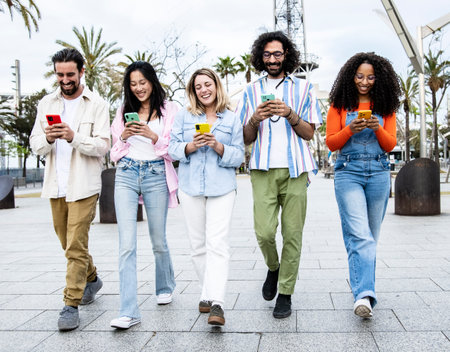 Multiethnic group of friends texting on their smartphones while walking in the street. Diverse young people smiling and using their phones outside in a city.の写真素材