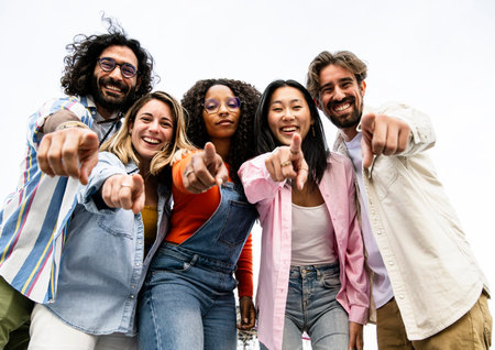 Low view of cheerful multiracial group of young hipsters pointing and looking at camera. Happy diverse group of young hipsters staring at camera outside and pointing fingers at the camera.の写真素材