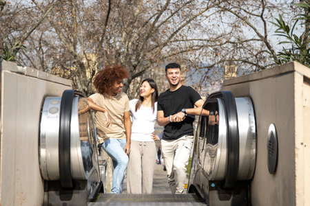 Diverse group of friends going up an outdoor escalator in a park. Three happy young people smiling and looking relaxed climbing up the stairs in the street.の写真素材