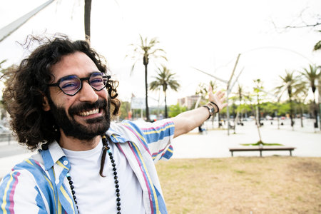 Portrait of cheerful happy hipster young man smiling looking at camera taking a selfie in the street with one arm wide open. Joyful carefree young guy laughing and staring at camera in the city.の写真素材