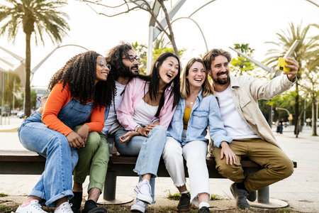 Diverse group of friends taking a selfie in the street sitting on a bench. Cheerful multiracial group of young hipsters taking a picture outside in a park bench.の写真素材