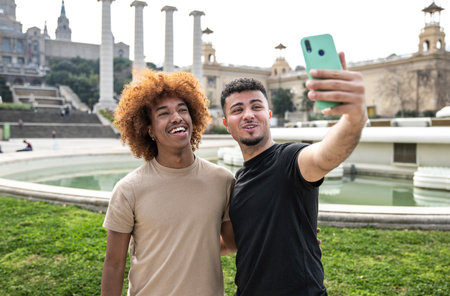 Two happy men taking a selfie with phone in park. Cheerful masculine couple smiling and taking a picture outside.の写真素材