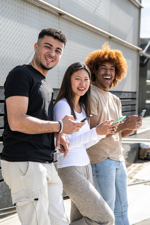 Three young diverse people laughing using smartphone and looking at camera. Multiracial group having fun and using phones standing in the streetの写真素材