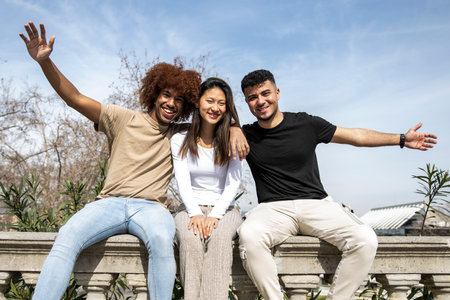 Multiracial group of young smiling people sitting on a stone balcony. Three diverse happy friends having fun together in a park.の写真素材