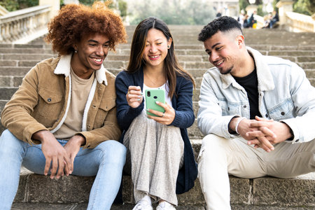 Three young diverse people looking at a smartphone sitting on the stairs.Two young men smiling and watching at the phone of a happy woman.の写真素材