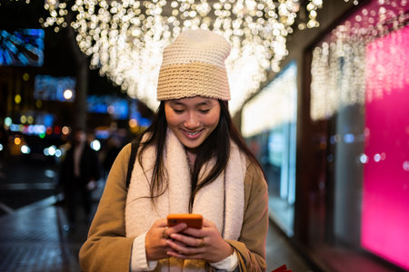 Young smiling girl texting in the street at night. Happy woman wearing warm hat using her phone in winter.の写真素材