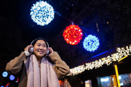 Young happy woman wearing headphones with christmas lights behind. Joyful girl listening to music at the street during winter.の写真素材