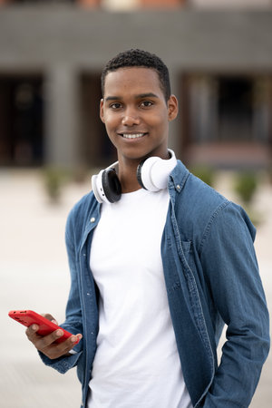 Young handsome guy smiling and looking at camera relaxed and using his phone.Satisfied man smiling and standing and texting while wearing headphones.の写真素材
