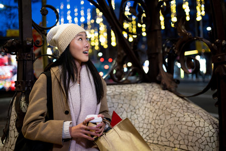 Joyful young woman shopping at evening staring at beautiful lights. Expressive female holding a coffee looking up at night in the street.の写真素材