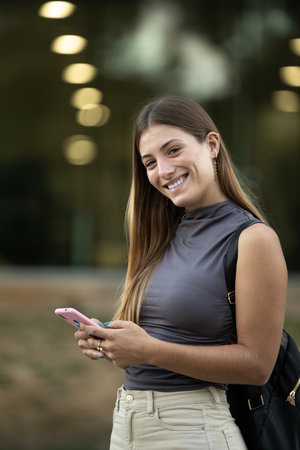 Beautiful cheerful young lady looking at camera texting and standing outside. Joyful blond woman staring at camera in the street while using her phone.の写真素材