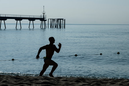 Running man silhouette training at the beach. Athlete jogging next to the sea with a sea dock in background.の写真素材