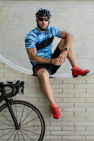 Bike rider in sport clothes sitting in a wall with a cycle looking at camera. Fit cyclist resting next to his cycle wearing helmet and sunglasses.の写真素材