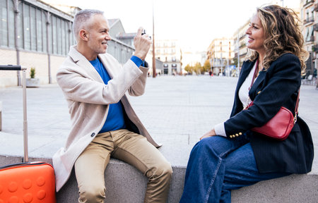 Senior tourist couple taking picture on vintage camera sitting in a bench. Mid adult man taking a...