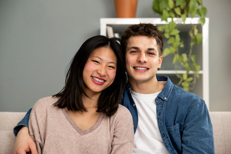 Affectionate smiling young adult couple sitting on sofa at home looking at camera. Carefree man and woman relaxing together in couch at the living room.の写真素材