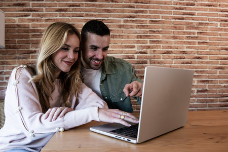 Young smiling couple connected on internet with laptop at their home. Joyful man and woman using...