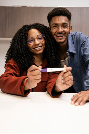 Affectionate happy young couple showing positive pregnancy test and hugging at home. Handsome young adult man holding maternity test with his wife embracing looking at camera.の写真素材