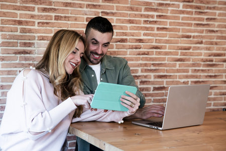 Happy man and woman using laptop and tablet sitting on a modern setting. Young adult couple working together with laptop from home in a modern apartment.の写真素材