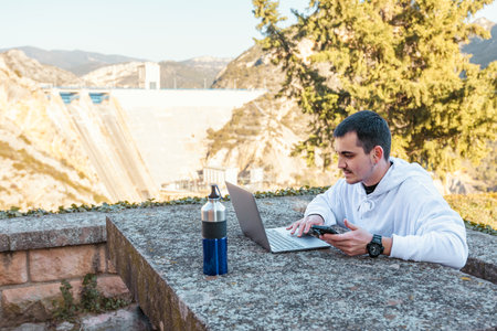 young digital nomad boy working outdoors with a laptop and mobile phone, while enjoying a warm and cozy landscapeの写真素材