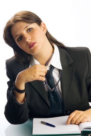 a young woman in black jacket and white shirt and a tie sitting down behind an office table and playing with glassesの写真素材