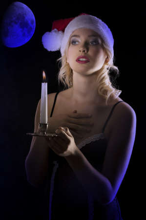 cute and attractive young woman with nightgown ant a christmas hat looking in the dark with a candle の写真素材